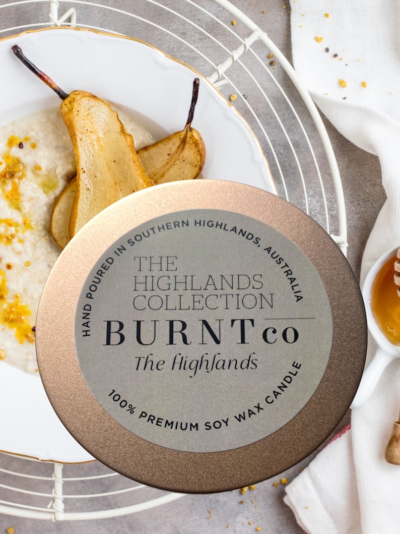 Puede incluir: Una lata de vela redonda y met&aacute;lica con el texto "BURNTco The Highlands" est&aacute; centrada en la imagen. La vela est&aacute; rodeada por un plato blanco con peras cocidas y un plato blanco con miel.