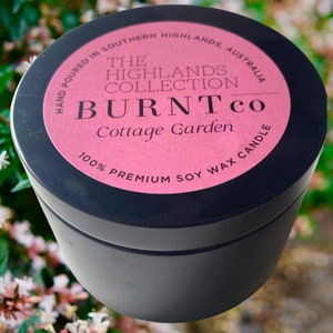 Puede incluir: Un recipiente negro para velas con una etiqueta rosa. La etiqueta dice "BURNT co Cottage Garden" y "100% Premium Soy Wax Candle". Tambi&eacute;n es visible el texto "Hand poured in Southern Highlands, Australia". La vela est&aacute; sobre un fondo floral borroso.