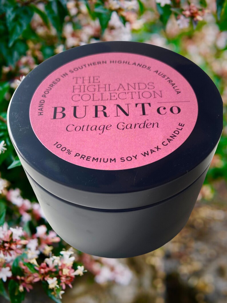 Puede incluir: Un recipiente negro para velas con una etiqueta rosa. La etiqueta dice "BURNT co Cottage Garden" y "100% Premium Soy Wax Candle". Tambi&eacute;n es visible el texto "Hand poured in Southern Highlands, Australia". La vela est&aacute; sobre un fondo floral borroso.