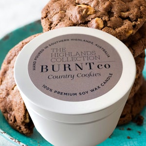 Puede incluir: Una vela blanca con la etiqueta "BURNT co Country Cookies" est&aacute; sobre un plato turquesa junto a galletas con chispas de chocolate. La vela est&aacute; etiquetada como "100% Premium Soy Wax Candle". Las galletas son marrones con nueces visibles.