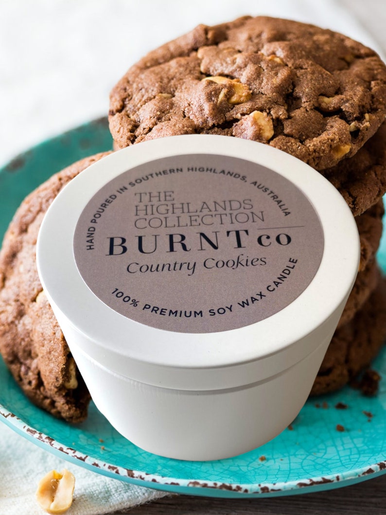 Puede incluir: Una vela blanca con la etiqueta "BURNT co Country Cookies" est&aacute; sobre un plato turquesa junto a galletas con chispas de chocolate. La vela est&aacute; etiquetada como "100% Premium Soy Wax Candle". Las galletas son marrones con nueces visibles.