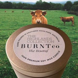 Puede incluir: Un bote de vela redondo, con borde dorado, que muestra el texto "BURNT Co" y "The Bowraf" en su etiqueta. La etiqueta tambi&eacute;n indica "100% Premium Soy Wax Candle". El fondo muestra un campo verde con vacas.
