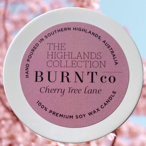 Puede incluir: Un recipiente blanco para velas con una etiqueta rosa que dice "BURNT co Cherry Tree Lane" y "100% Premium Soy Wax Candle". La etiqueta tambi&eacute;n indica "Hand Poured in Southern Highlands, Australia". El fondo es un desenfoque suave de flores rosas.