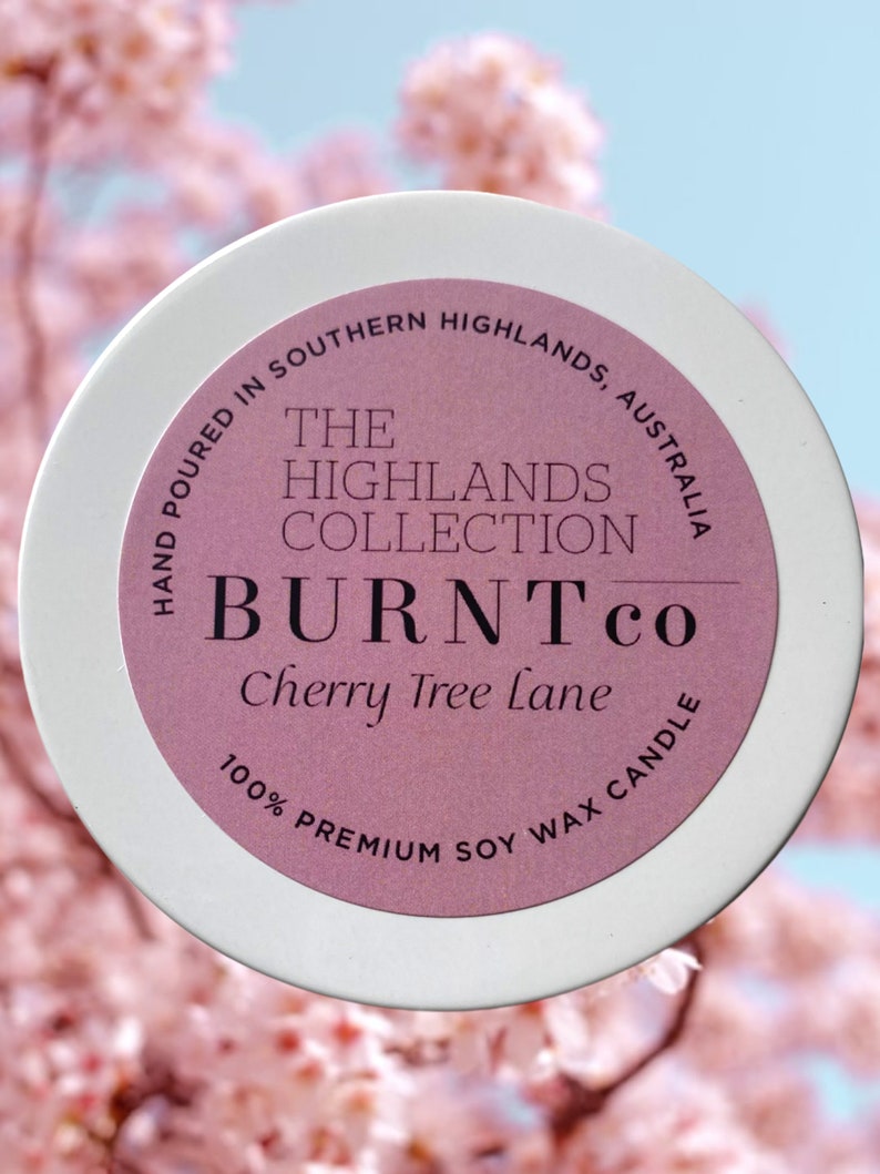 Puede incluir: Un recipiente blanco para velas con una etiqueta rosa que dice "BURNT co Cherry Tree Lane" y "100% Premium Soy Wax Candle". La etiqueta tambi&eacute;n indica "Hand Poured in Southern Highlands, Australia". El fondo es un desenfoque suave de flores rosas.