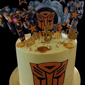 Pode incluir: Um bolo redondo, com cobertura amarela, decorado com gráficos Transformers laranja e preto. O bolo é coberto com recortes de personagens Transformers, decorações em forma de estrela, moedas de ouro e o número 5. O bolo está sobre uma base dourada.