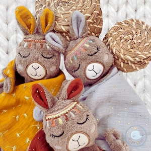 May include: Three brown plush llama toys with embroidered eyes and a colorful beaded headband. The llamas are lying on a yellow and gray blanket with gold glitter.