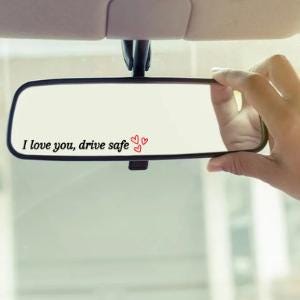 May include: A rectangular rearview mirror with the text "I love you, drive safe" and three red hearts. The mirror is held by a hand, with the car's interior visible in the background.