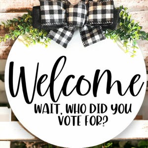 Op de afbeelding: Een ronde witte houten plank met het woord "Welcome" in zwart schrift. Daaronder staat de tekst "WAIT, WHO DID YOU VOTE FOR?". Het bord is versierd met een zwart-wit geruite strik en groen.