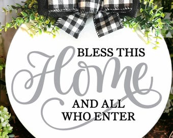May include: A circular white wooden sign featuring the words "BLESS THIS Home AND ALL WHO ENTER" in grey script. It's adorned with a black and white check bow and green foliage, ideal for home decoration.