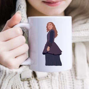 May include: White ceramic mug with a stylized illustration of a person in a dark purple dress and jacket. The figure has long auburn hair. The mug is held by a person wearing a cream-coloured sweater.