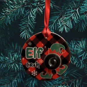 May include: Round Christmas ornament with a red and black plaid pattern. The ornament features the text "Elf Cam" in green and white, along with a camera lens and elf hat graphics. A red ribbon is attached for hanging.
