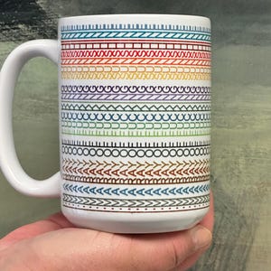 May include: White ceramic mug with a large handle, featuring a repeating pattern of colorful geometric designs in a rainbow of colors. The mug is held in a person's hand.