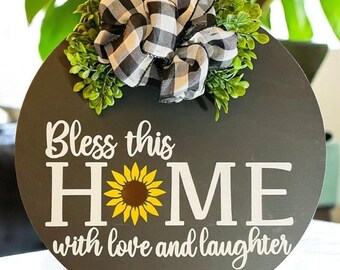 May include: A round wooden sign with a grey background and white lettering that reads "Bless this HOME with love and laughter". A sunflower graphic is in the centre of the word "HOME". A black and white checked bow with green foliage is at the top of the sign.