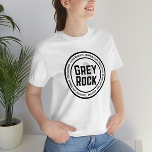 May include: White t-shirt with a black and grey circle graphic that says "Grey Rock" and has the words "Housewarming, Gaslighting, Love Bombing, Scapegoating, Flying Monkeys, Triangulation, Projection, Blame Shifting" around the circle.