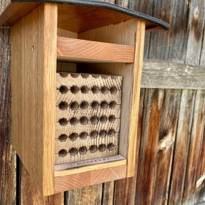 May include: A wooden bee house featuring a light brown frame and a dark roof. The interior contains a block of wood with a grid of circular holes drilled into the front surface, providing nesting tunnels for solitary bees in a natural wood finish.