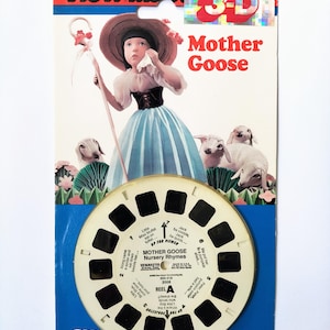 Puede incluir: Un carrete View-Master 3D con rimas infantiles de Mother Goose. El carrete es blanco con imágenes y texto negros. La parte delantera del embalaje presenta una ilustración de dibujos animados de una niña con un vestido azul y un sombrero de paja, sosteniendo un cayado de pastor. Está rodeada de ovejas blancas. El texto "Mother Goose" está en letras rojas grandes.