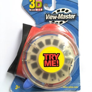 May include: A red and blue View-Master virtual 3D viewer with a clear plastic lens and a "Try Me!" sticker. The viewer is packaged in a blue and white cardboard box with the View-Master logo.
