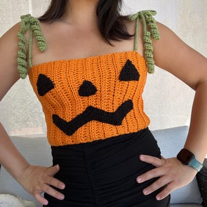 Puede incluir: Un top de calabaza de color naranja tejido a crochet con rasgos faciales negros y tirantes de crochet verde. El top lo lleva una persona con una falda negra.