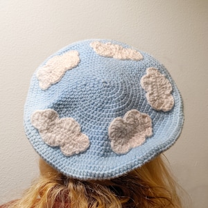 May include: A light blue crocheted beret with white cloud appliques. The beret is worn on a person's head.
