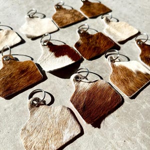 May include: A collection of cowhide keyrings with silver key rings. The keyrings are shaped like a cow tag and have a brown and white pattern.