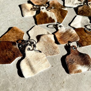 May include: A collection of brown and white cowhide keyrings with silver key rings. The keyrings are shaped like a cow tag.