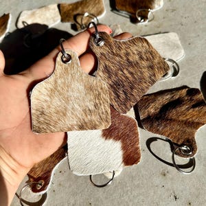 May include: A set of cowhide keyrings with brown and white patterns. Each keyring features a metal ring and a leather tag shaped like a cow's ear tag.