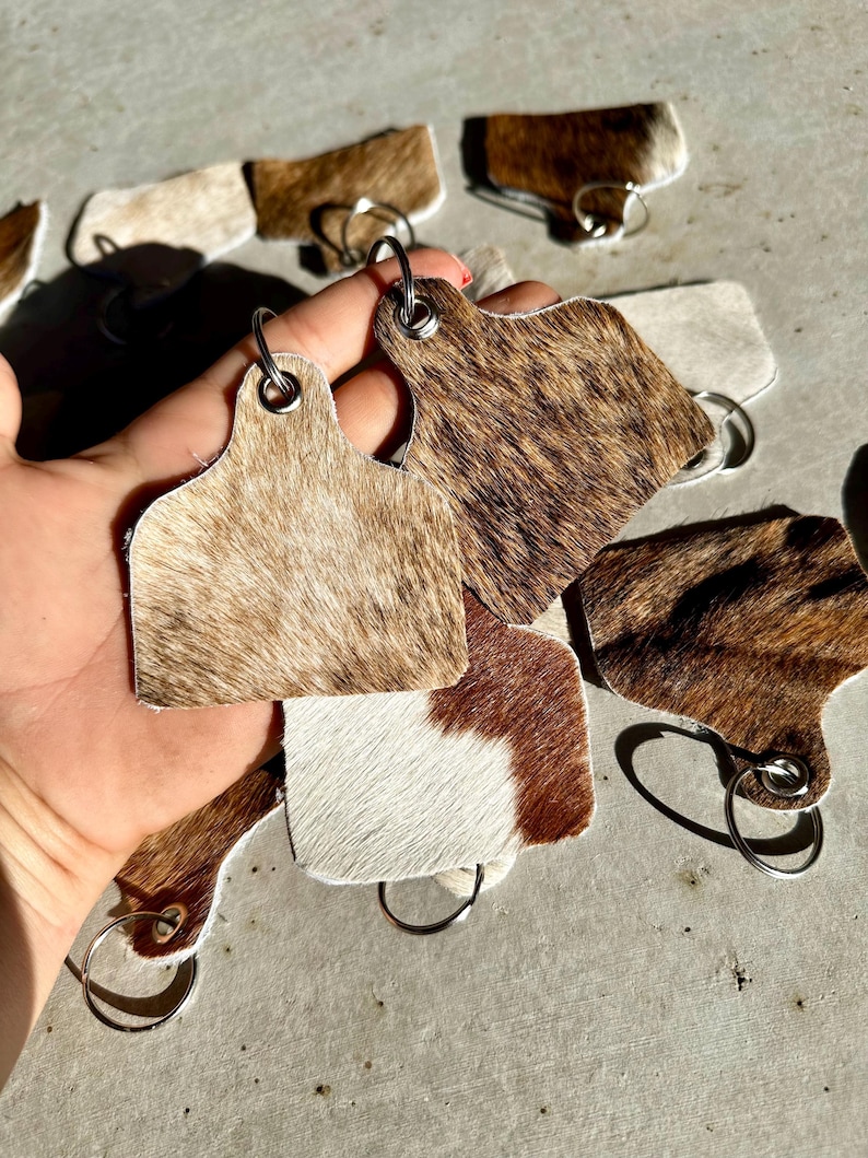 May include: A set of cowhide keyrings with brown and white patterns. Each keyring features a metal ring and a leather tag shaped like a cow's ear tag.
