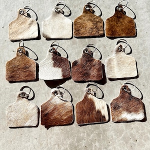 May include: Twelve cowhide keyrings with silver key rings. The keyrings are shaped like a cow tag and have a brown and white pattern.