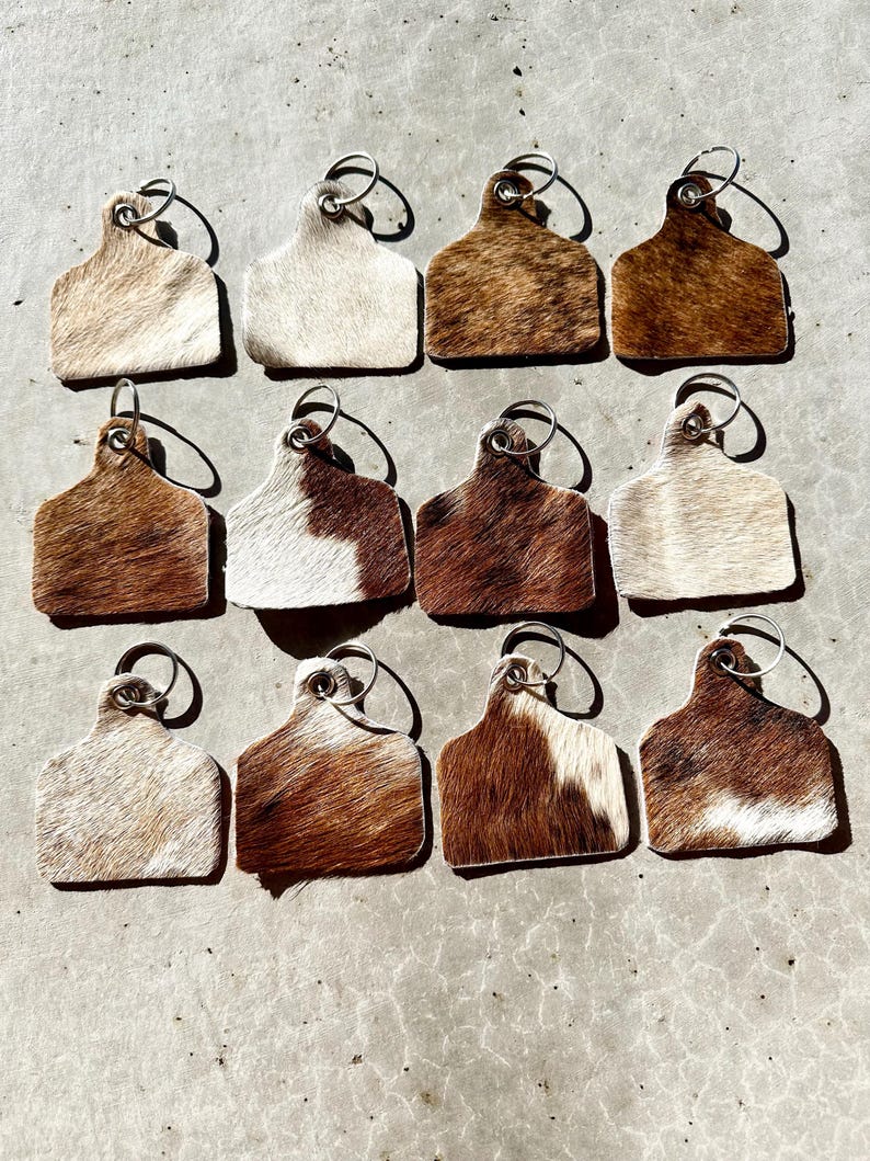 May include: Twelve cowhide keyrings with silver key rings. The keyrings are shaped like a cow tag and have a brown and white pattern.
