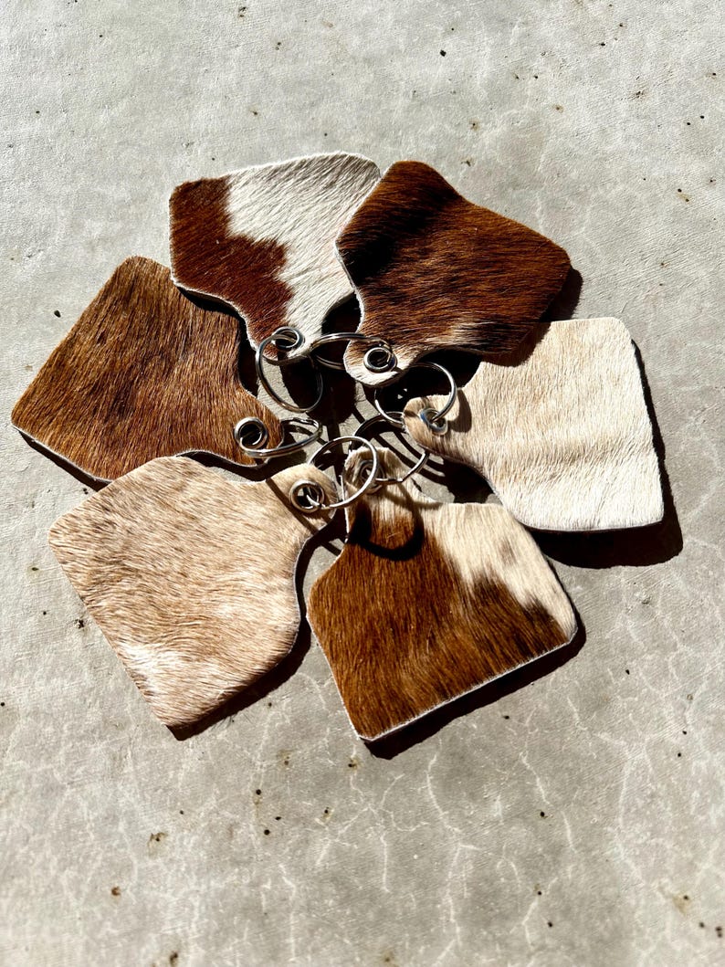May include: Six brown and white cowhide keyrings with silver key rings. The keyrings are shaped like a cow tag.