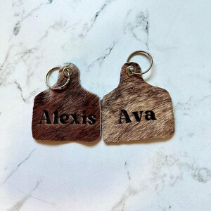 May include: Two brown leather keychains with silver key rings. The keychains are shaped like cow tags and have the names "Alexis" and "Ava" embossed on them.