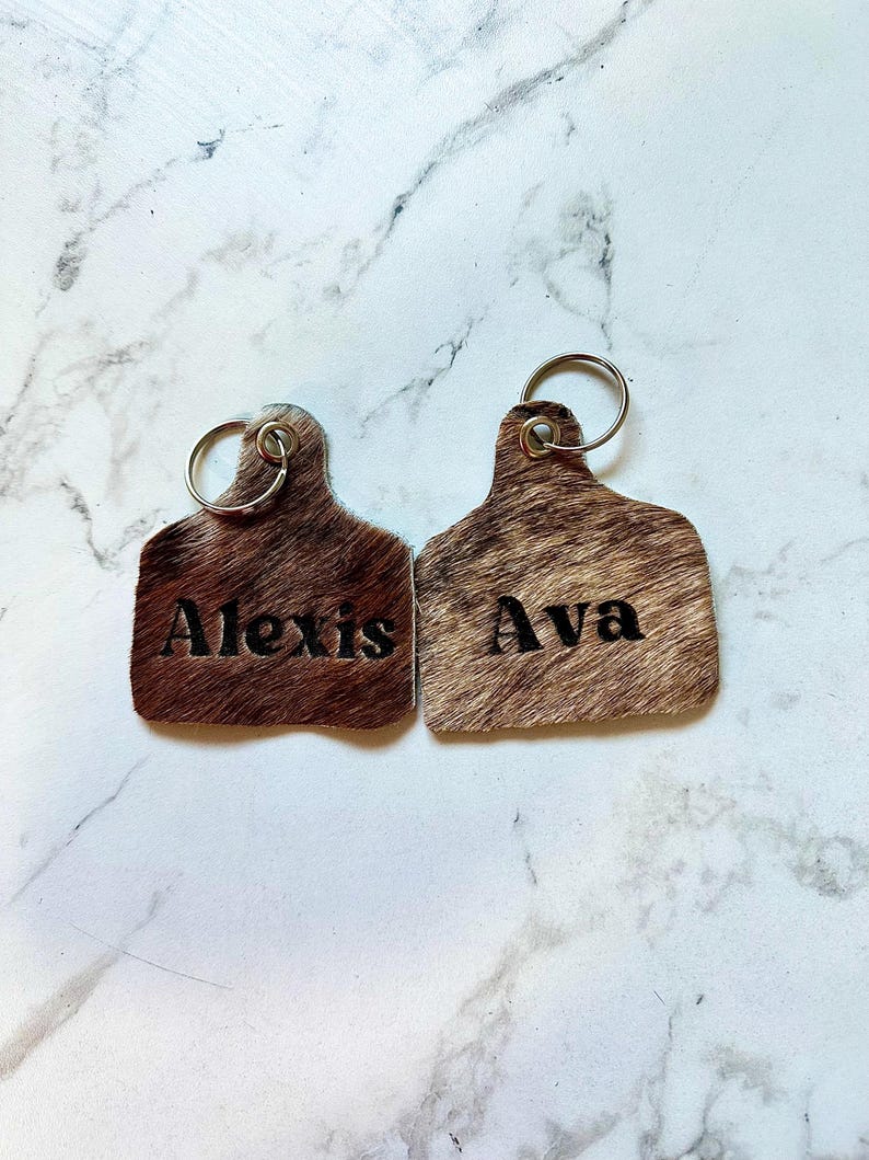 May include: Two brown leather keychains with silver key rings. The keychains are shaped like cow tags and have the names "Alexis" and "Ava" embossed on them.