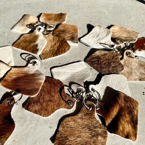 May include: A set of cowhide keyrings with silver key rings. The keyrings are in various shades of brown and white.
