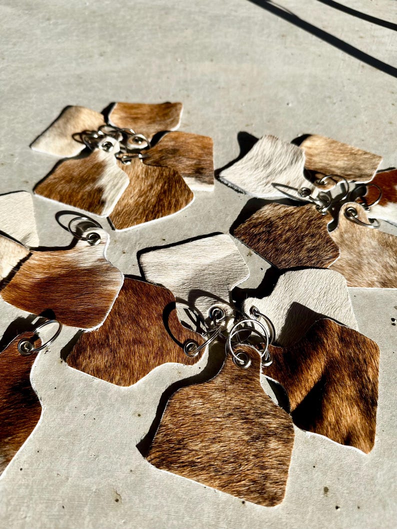 May include: A set of cowhide keyrings with silver key rings. The keyrings are in various shades of brown and white.