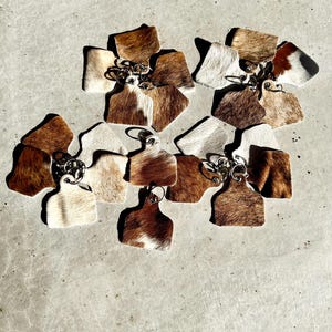 May include: A group of ten cowhide keyrings in various shades of brown and white. The keyrings are shaped like a cow tag and have a metal ring attached.