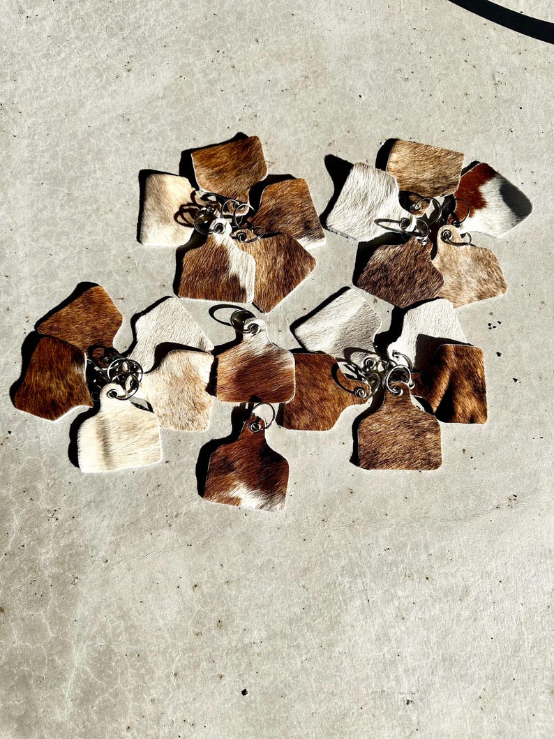 May include: A group of ten cowhide keyrings in various shades of brown and white. The keyrings are shaped like a cow tag and have a metal ring attached.