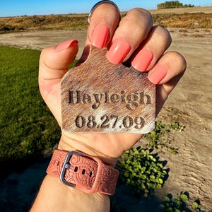 May include: A brown leather keychain with the name "Hayleigh" and the date "08.27.09" engraved on it. The keychain is being held by a hand with pink nail polish.