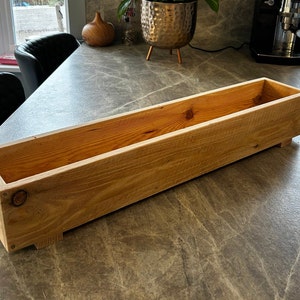 May include: A long, rectangular wooden planter box with a natural wood finish. The planter has a simple design with no embellishments. It is made of unfinished wood and has a rustic look.