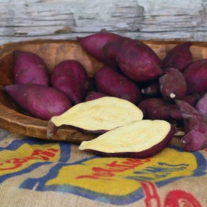 May include: A wooden bowl filled with vibrant purple sweet potatoes, with a few cut open to reveal the pale yellow flesh. The potatoes rest on a burlap sack with colorful text.