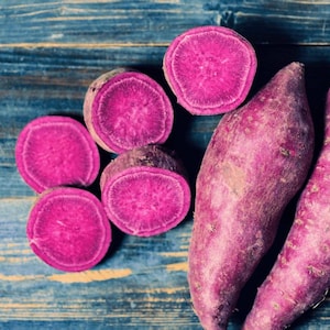 May include: Overhead shot of purple sweet potatoes, some whole and some sliced, revealing a vibrant magenta interior. The potatoes rest on a weathered, blue-painted wooden surface, creating a rustic and colorful composition. A healthy food concept.