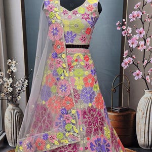 May include: A sleeveless beige lehenga choli set featuring a vibrant, multi-colored floral pattern in neon pink, yellow, purple, and orange. The ensemble includes a matching sheer dupatta with embroidered floral borders and intricate sequin detailing.