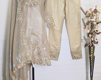 May include: A beige three-piece outfit consisting of a long-sleeved tunic, matching trousers, and a dupatta. The tunic features intricate gold embroidery, beadwork, and mirror accents around the neckline and hem. The trousers have embroidered cuffs.