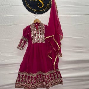 May include: A magenta-colored dress with intricate gold embroidery and embellishments. The dress features long sleeves, a high collar, and a flowing skirt. A matching magenta scarf with gold trim is draped to the side. The dress is hanging on a wooden hanger.