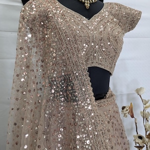 Może przedstawiać: Beżowy zestaw lehenga choli z cekinami, wystawiony na manekinie. Strój posiada skomplikowane hafty i pasującą dupattę. Złoty naszyjnik zdobi szyję manekina. Tło to gładka biała powierzchnia.