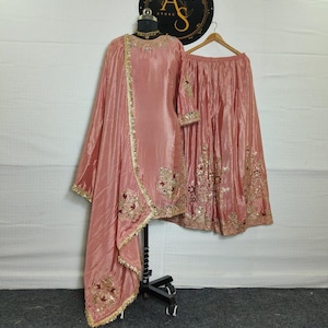 May include: A blush pink Indian salwar kameez set. The set includes a long tunic, a skirt, and a shawl. The tunic and skirt are embellished with gold embroidery and sequins. The shawl has a gold border. The outfit is displayed on a mannequin.