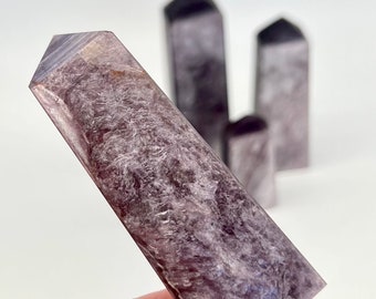 May include: A purple and grey crystal point, also known as a wand, is held in a hand. The crystal is smooth and has a pointed tip.