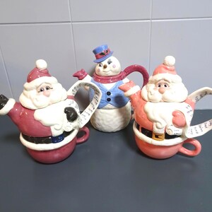 May include: Three ceramic Christmas teapots: two Santa Claus and one snowman. The Santa teapots are red and white with black gloves and belts. The snowman has a blue top hat and vest. The teapots are on a grey surface.