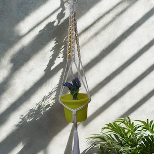 May include: A hanging macrame plant holder with a yellow pot containing a small plant with purple flowers. The hanger is white and yellow, suspended from a metal ring. The background is a textured wall with shadows.