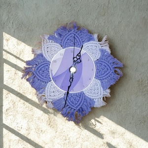 May include: A decorative wall clock with a macrame design in shades of lavender and white. The clock face is a gradient of purple hues, with black clock hands. The clock is mounted on a textured, light-colored wall.