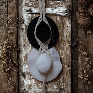 May include: A macramé hat holder with a wooden ring at the top, holding a black hat and a white hat. The hats are suspended against a weathered, rustic wooden door. The macramé is beige, and the hats have wide brims.